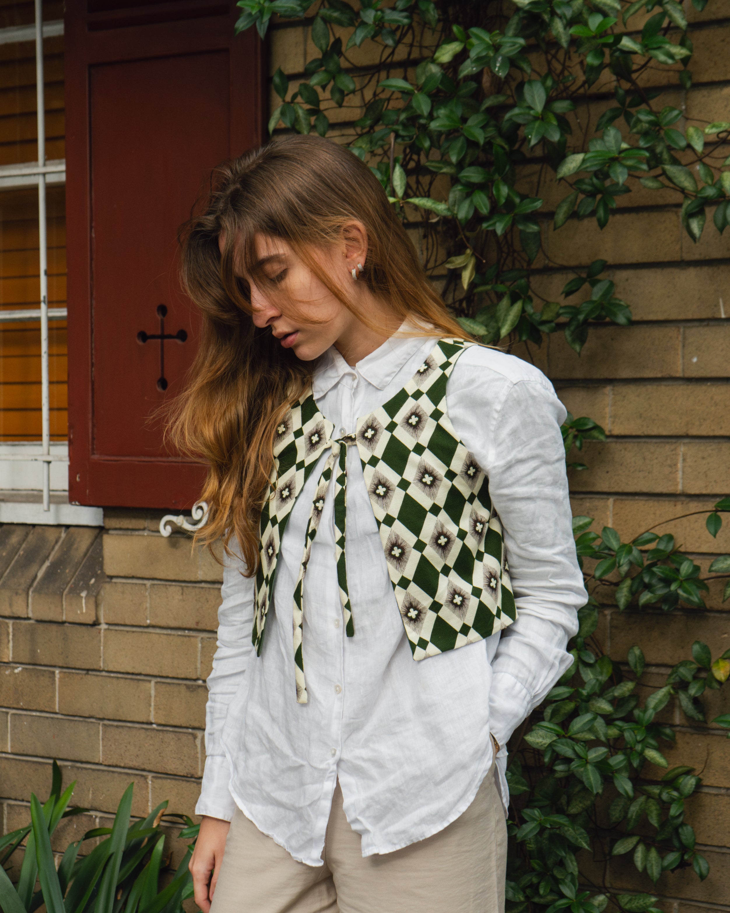 Woman wearing an Australian handmade green vest with a white shirt underneath of it in front of a brick wall with leaves